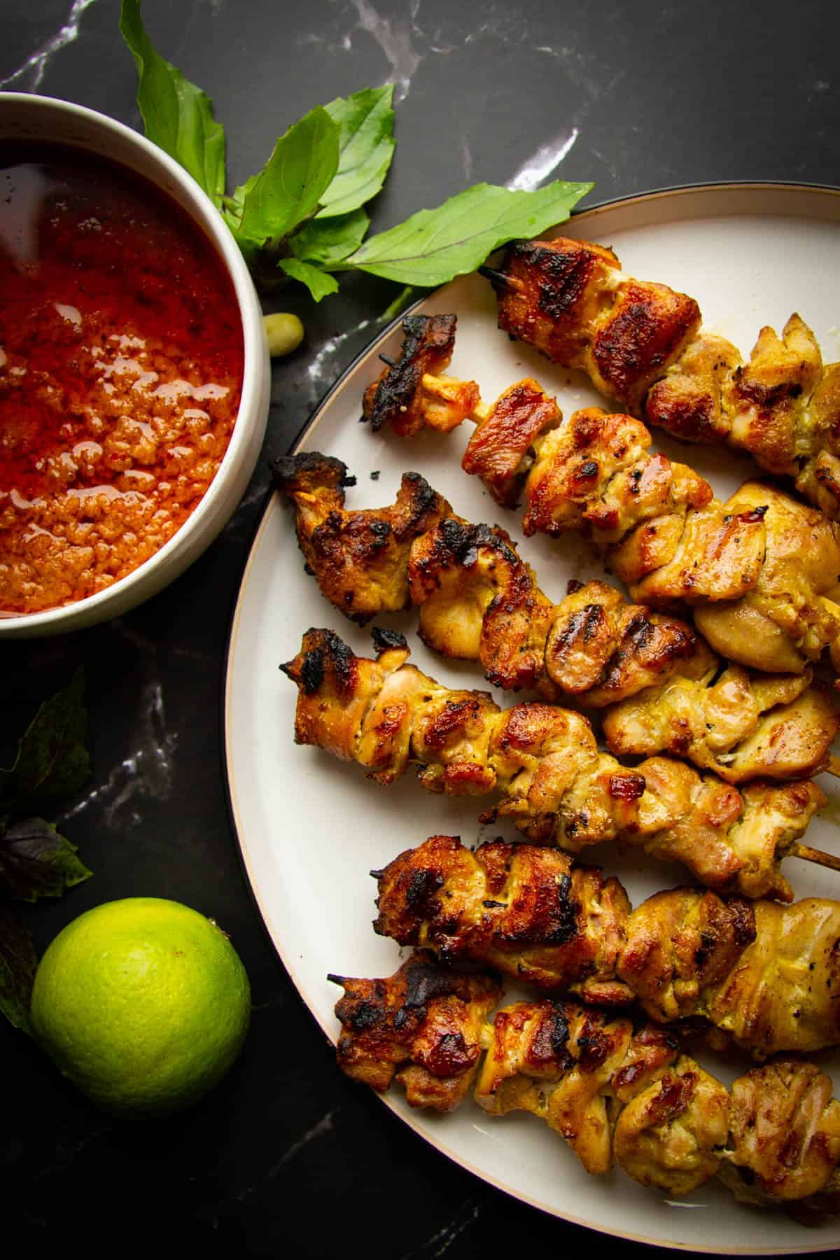charcoal chicken satays on a plate with peanut sauce, lime and basil on the side.