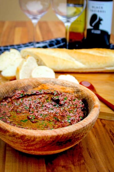 Olive tapenade in a wooden bowl with wine glasses and a sliced baguette beside.