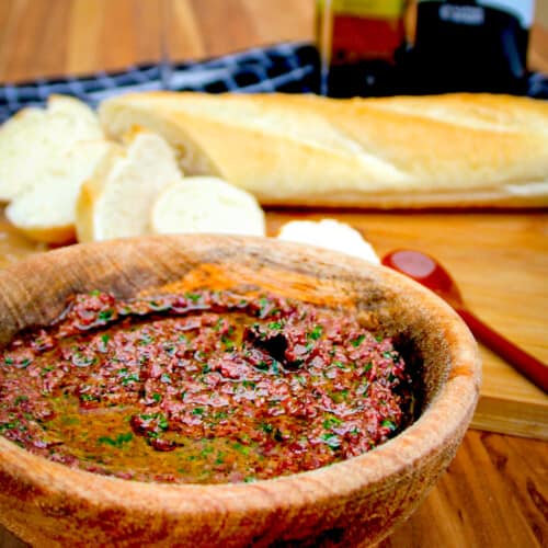 Olive tapenade in a wooden bowl with wine glasses and a sliced baguette beside.