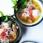 Pho in two bowls with fresh herbs, chili and bean sprouts on the side.