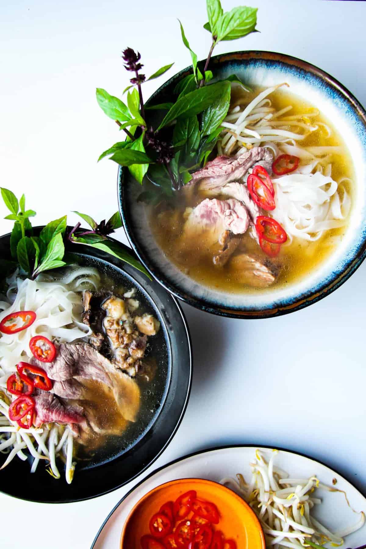 Pho in two bowls with fresh herbs, chili and bean sprouts on the side.