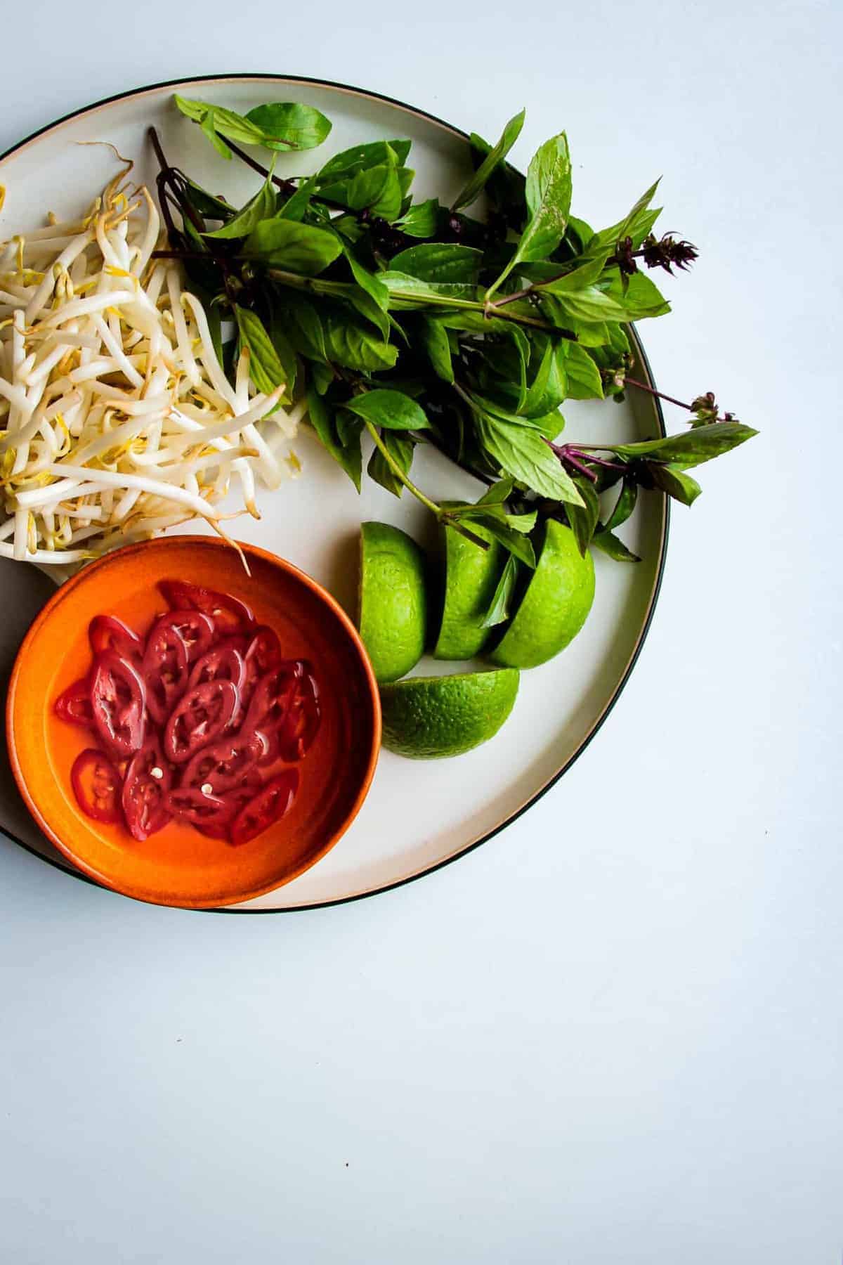 Fresh basil, sliced chili, bean sprouts and fresh lime wedges on a plate.