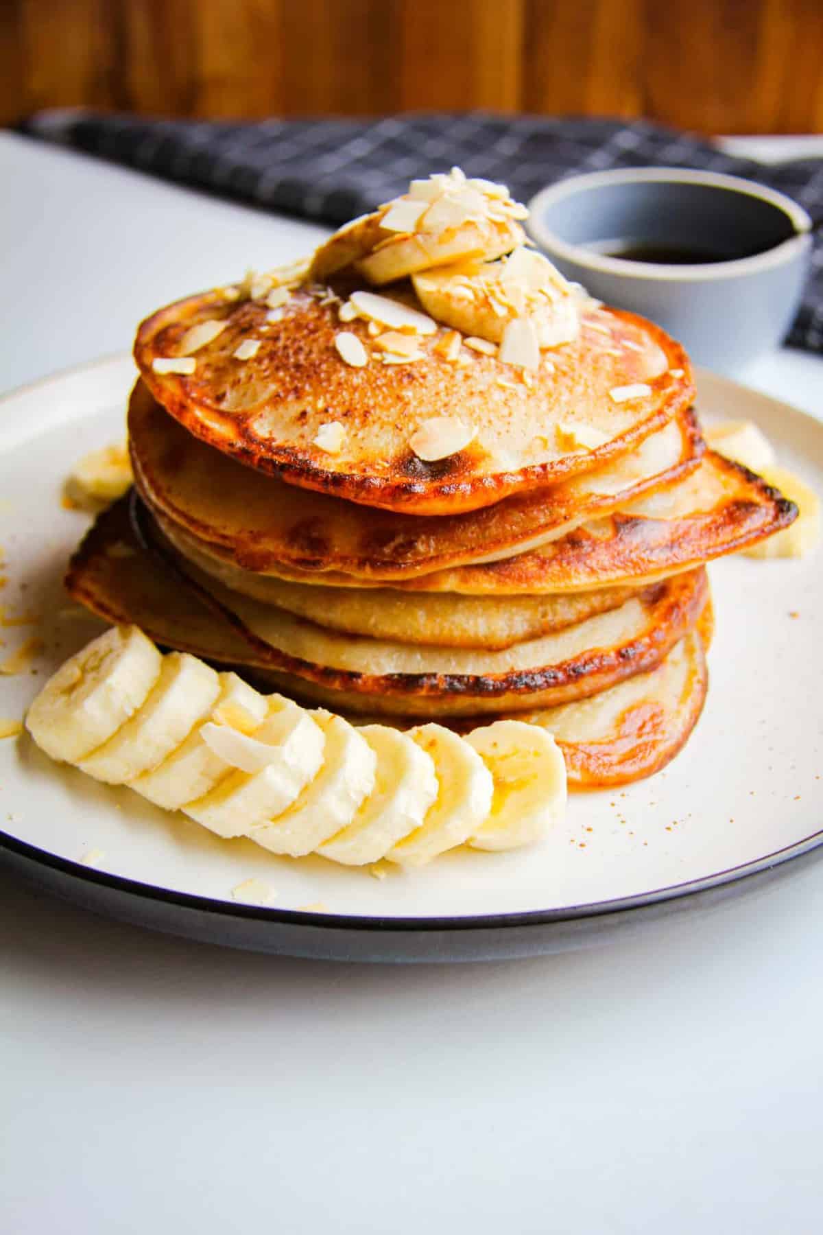 Vegan banana pancakes stacked high on a plate with almonds, fresh banana and maple syrup.