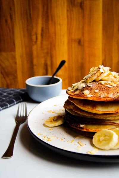 Vegan banana pancakes stacked high on a plate with almonds, fresh banana and maple syrup.