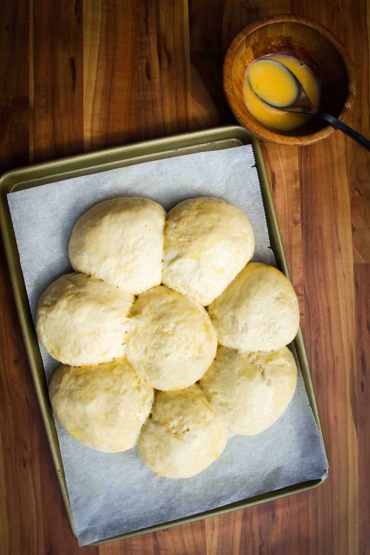 The dinner rolls proofing on a tray with egg wash on the side.