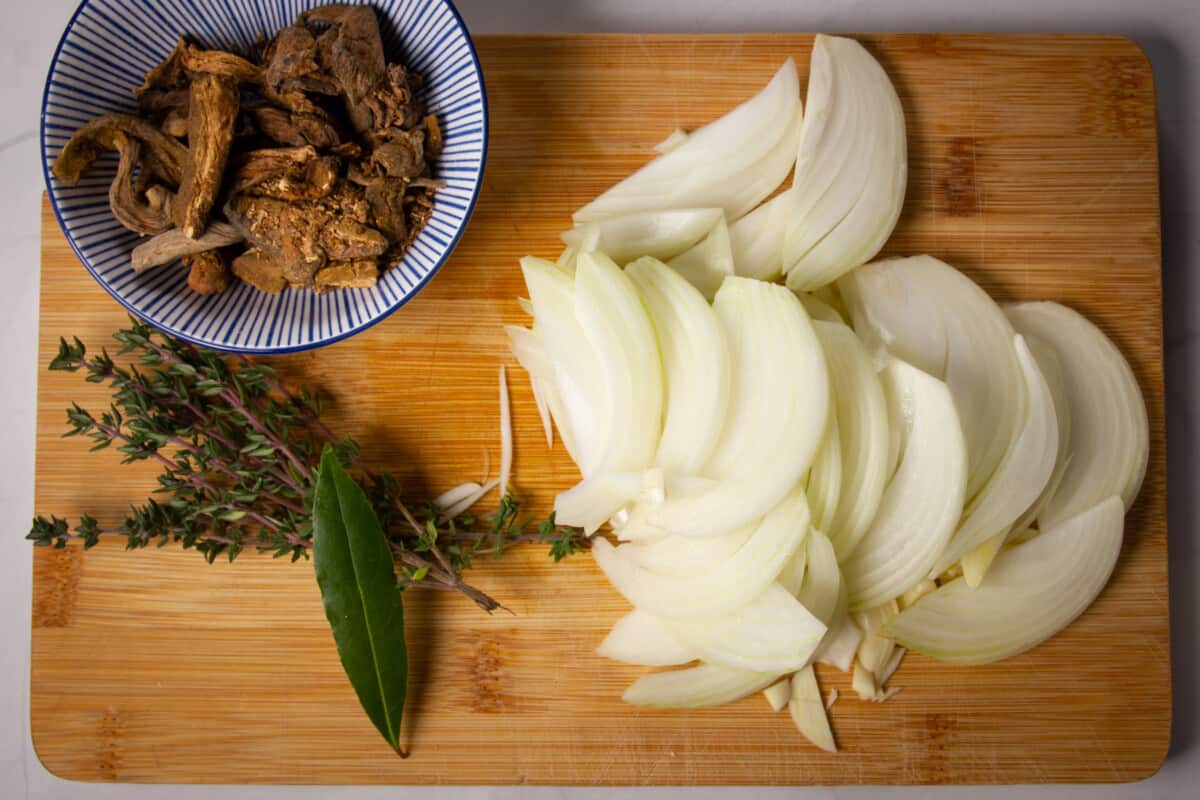 Sliced onions, garlic, herbs and dried mushrooms on a cutting board.