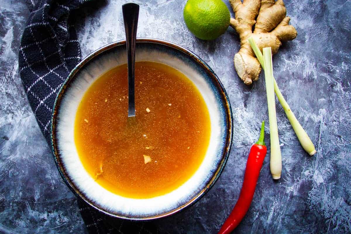 Instant Pot Chicken Bone Broth with Lemongrass + Chili in a bowl with a spoon and some chili, lemongrass and ginger beside it.