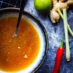 Instant Pot Chicken Bone Broth with Lemongrass + Chili in a bowl with a spoon and some chili, lemongrass and ginger beside it.