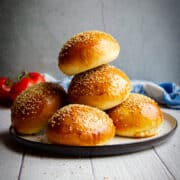 Super soft homemade hamburger buns stacked on a plate with tomatoes in the background.