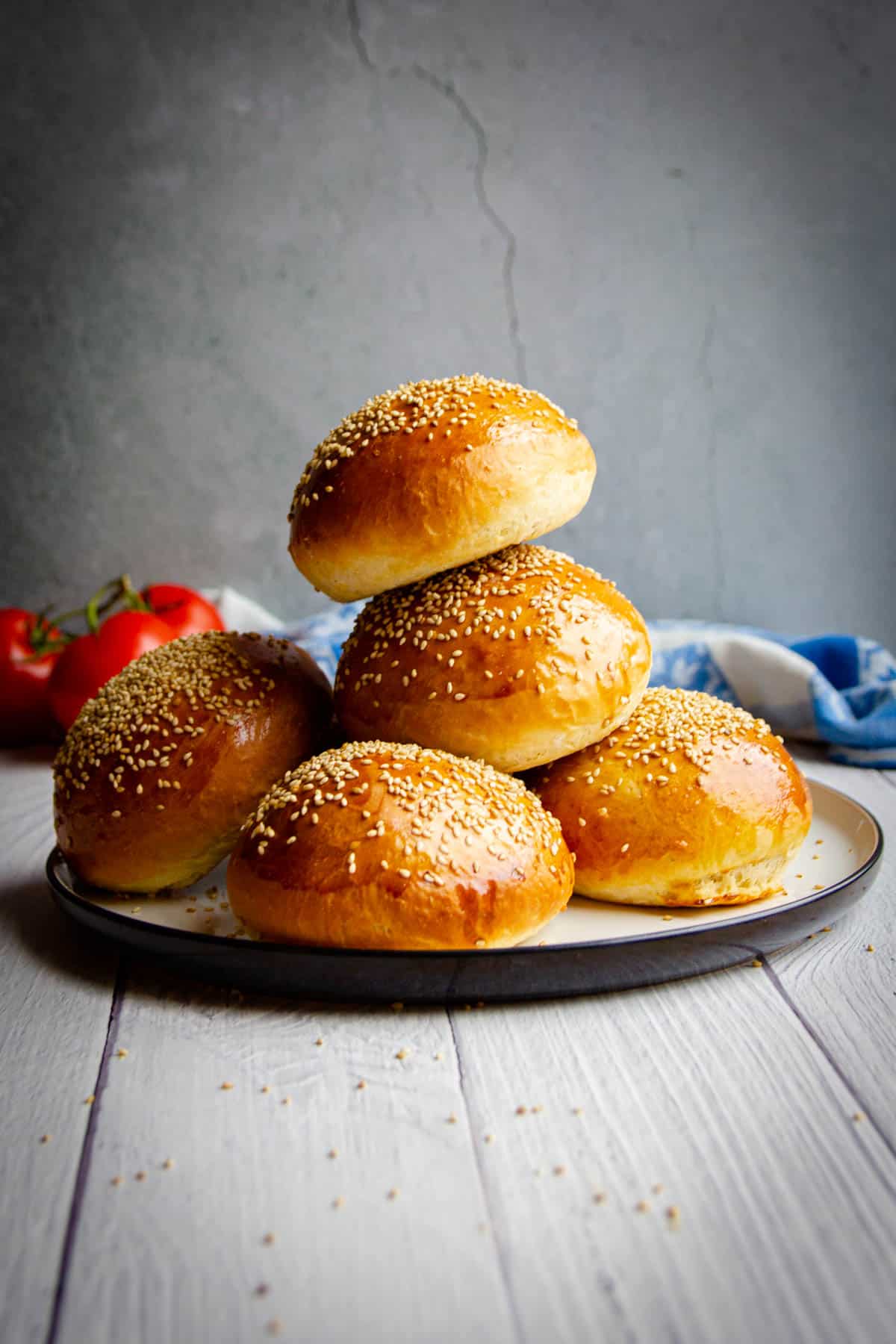 Super soft homemade hamburger buns stacked on a plate with tomatoes in the background.