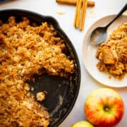 A slice of apple crisp on a plate with the rest of the apple crisp behind