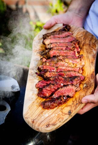 A smoked, reverse-seared ribeye sliced on a wooden board with smoke in the background.