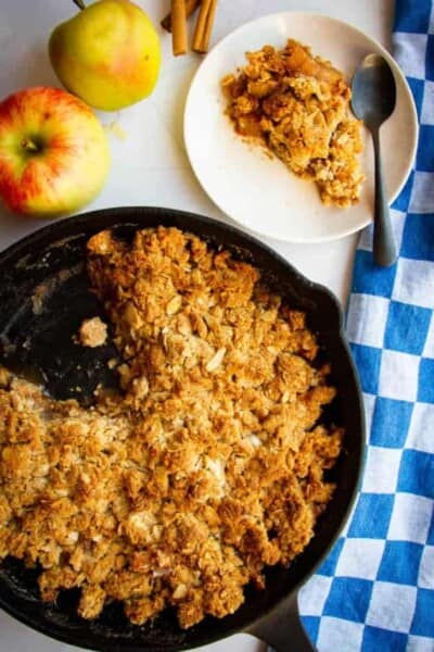 A cast iron pan full of apple crisp with apples in the background.