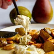 A fork scoopin into a piece of burrata on a plate with roasted pears, thyme, marcona almonds and brown butter.