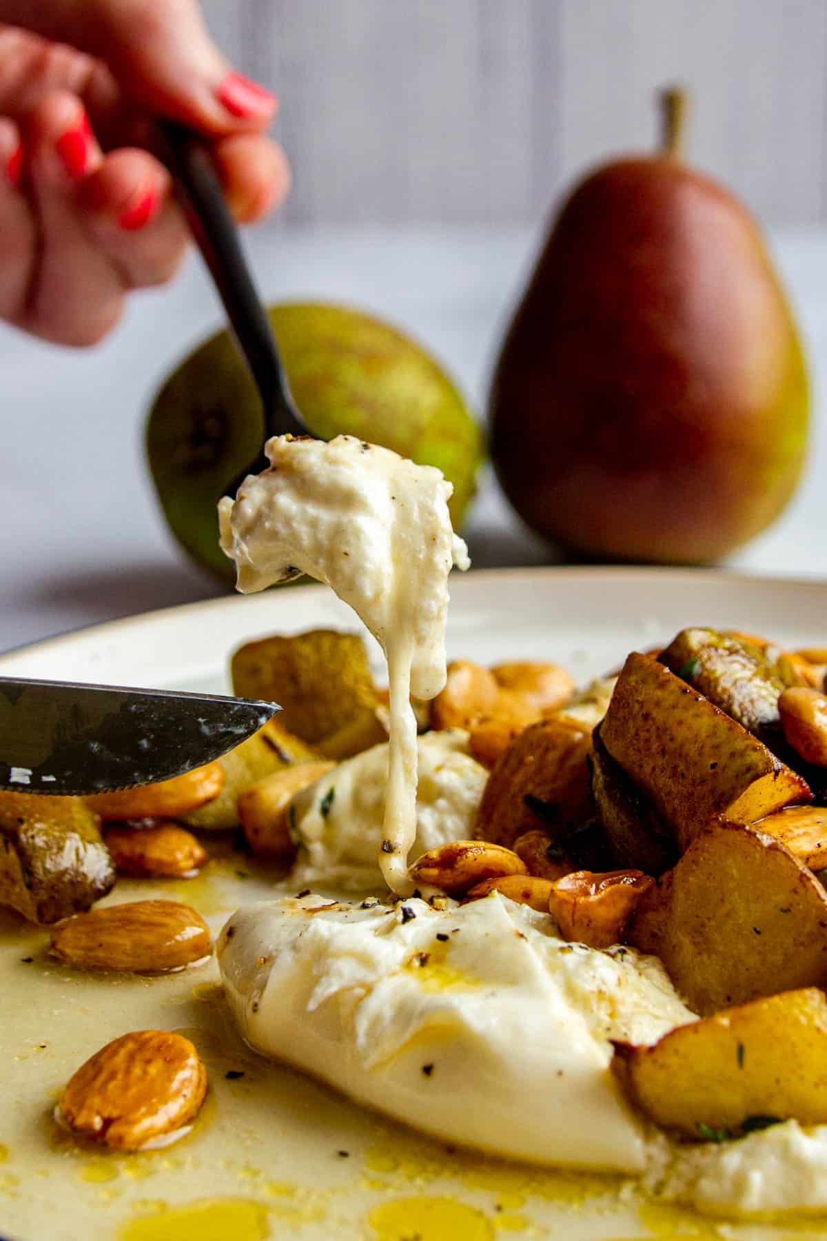 A fork scoopin into a piece of burrata on a plate with roasted pears, thyme, marcona almonds and brown butter.