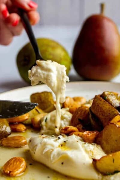 A fork scoopin into a piece of burrata on a plate with roasted pears, thyme, marcona almonds and brown butter.
