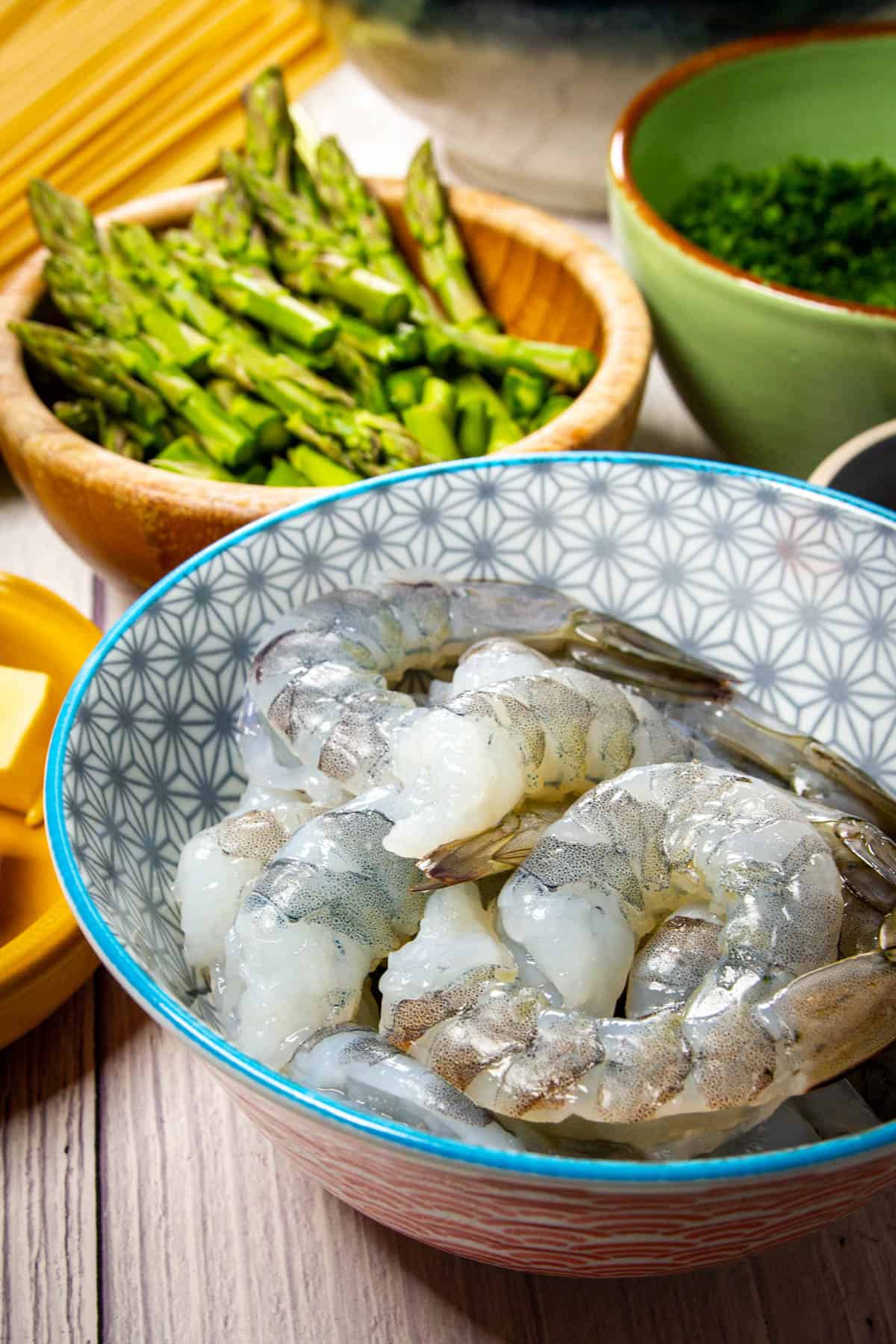 A bowl of raw shrimp with fresh asparagus in the background.