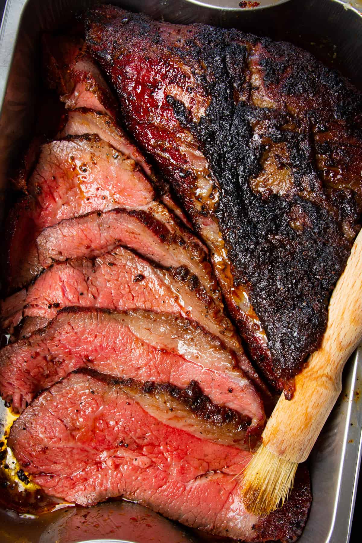 Smoked tri tip sliced up in a bbq tray with a brush beside it.